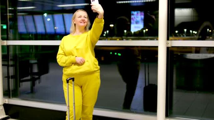 机场一名妇女手里拿着一个手提箱，拿着手机自拍亮黄色西装美丽的中年金发女郎晚上在街上靠近窗台期待一架飞