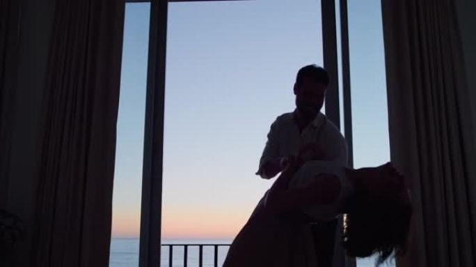 剪影快乐的夫妇在日落时在酒店房间跳舞，享受蜜月假期，享受美丽的海景庆祝假期