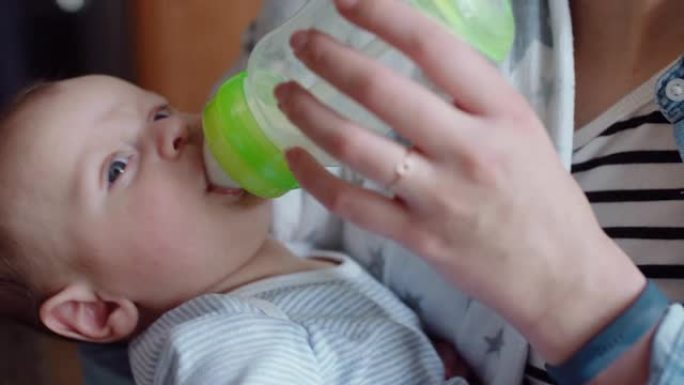 年轻的母亲喂养不快乐的婴儿从奶瓶中喝水，哭泣不安的婴儿在家与疲倦的妈妈