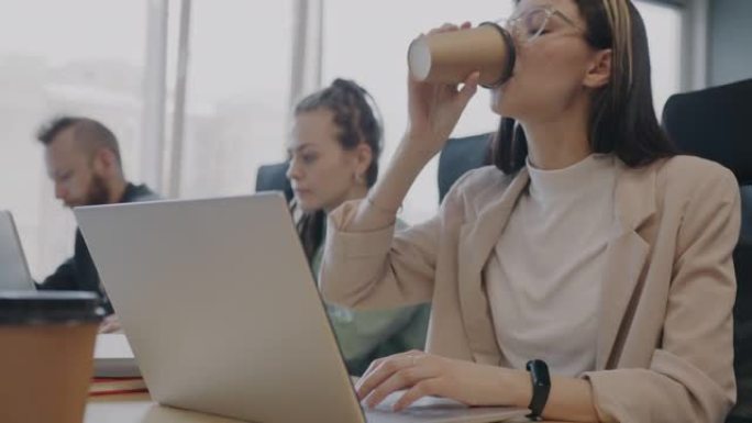 美丽的年轻女士使用笔记本电脑打字和喝酒去咖啡与同事一起在办公室工作
