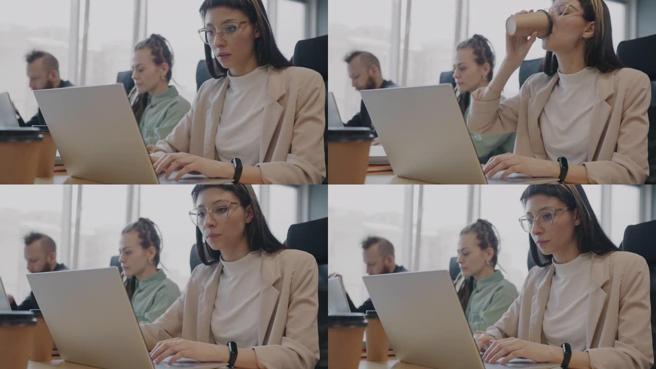 美丽的年轻女士使用笔记本电脑打字和喝酒去咖啡与同事一起在办公室工作