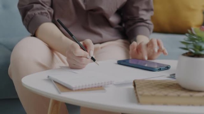 认真的年轻女子使用智能手机触摸屏并在笔记本上写字，专注于家里的生意