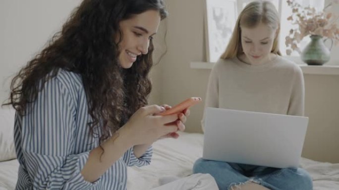 快乐的朋友使用小工具笔记本电脑和智能手机坐在床上，与电子设备聊天