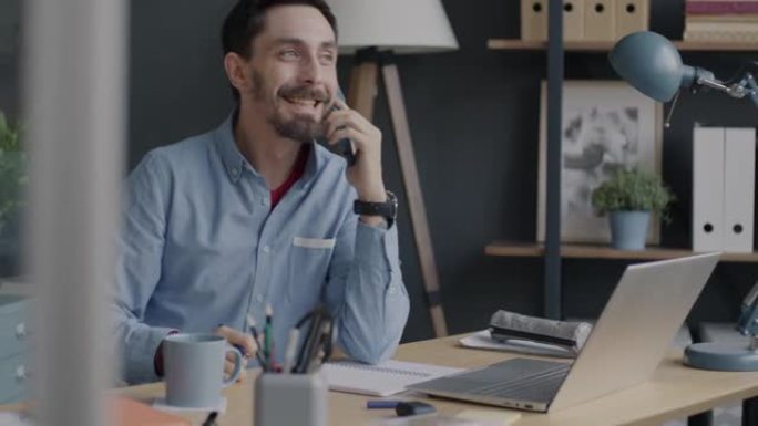 快乐的商人在手机上讲话并在笔记本上写信息，看着办公室的笔记本电脑屏幕