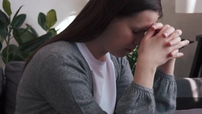 焦虑悲伤的年轻女子坐在家里的沙发上，担心困难的生活状况，健康问题或人际关系的困扰，迷失在消极的想法中