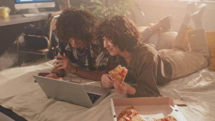 年轻夫妇躺在床上看笔记本电脑上的视频，吃披萨，早上