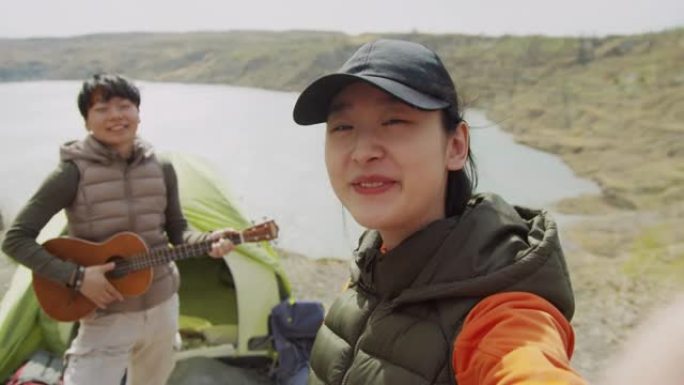 亚洲女性朋友在镜头前微笑着挥手，在湖边徒步旅行，一个带着吉他