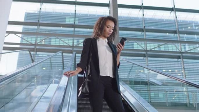 混血儿女商人在扶梯上使用智能手机发短信在线查看电子邮件成功的女高管离开公司办公室4k镜头