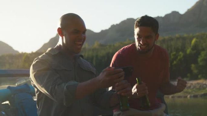 两个背着背包的男性朋友在湖边的卡车上喝啤酒在电话上观看体育比赛