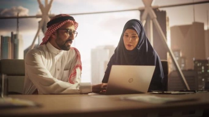 两名多元化的穆斯林财务顾问使用笔记本电脑在现代工业办公室讨论股票市场策略。阿拉伯男女经理在银行发展中