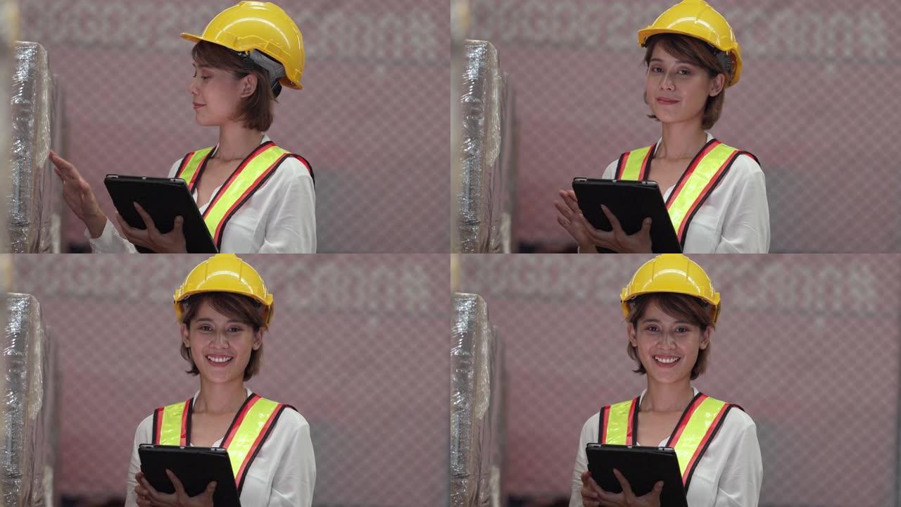 亚洲工人妇女拿着平板电脑在仓库检查库存。仓库工人穿着黄色反光安全背心和黄色头盔看着相机。