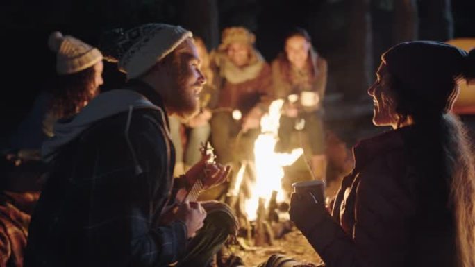 朋友们坐在篝火旁，年轻男子玩ukelele唱歌给晚上在森林里享受浪漫小夜曲露营的女人