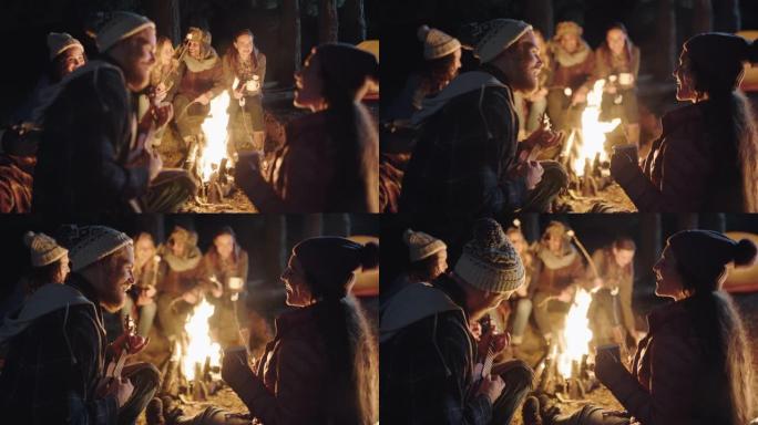 朋友们坐在篝火旁，年轻男子玩ukelele唱歌给晚上在森林里享受浪漫小夜曲露营的女人
