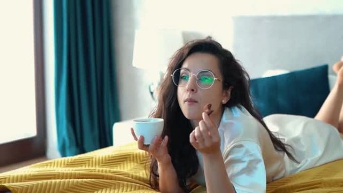 美丽的白人女性，戴着眼镜，躺在床上，饶有兴趣地看电视，用白色的杯子喝咖啡，吃饼干。女孩在卧室里吃巧克