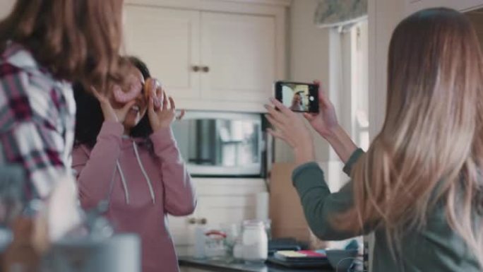 有趣的十几岁的女孩与甜甜圈合影，在社交媒体上分享智能手机，享受周末在厨房闲逛的乐趣