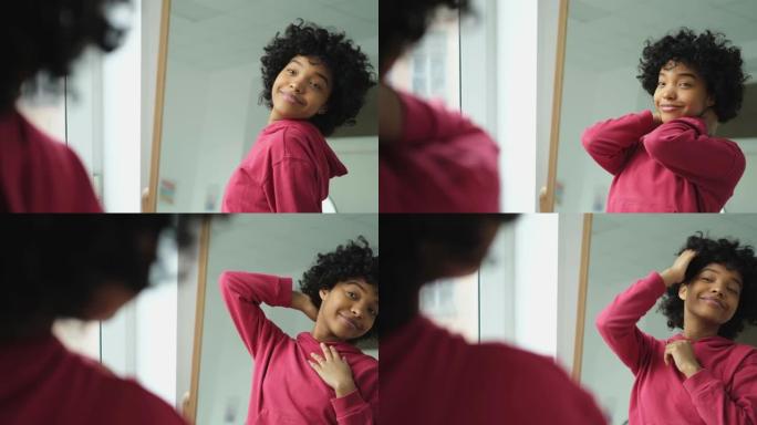 爱自己。美丽的年轻微笑的非洲裔美国妇女跳舞触摸卷发享受她的镜子反射。看着镜子的黑人女士看起来自信快乐