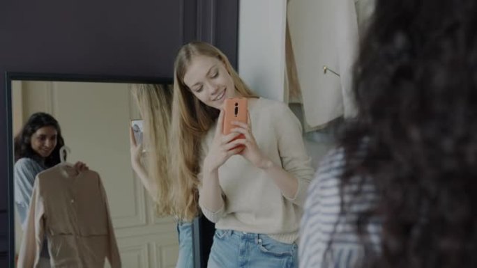 年轻女子拍摄朋友在商店里选择衣服的照片，为照镜子的智能手机相机摆姿势
