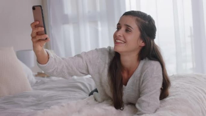 美丽的女人使用智能手机拍摄自拍照片，享受在社交媒体上分享生活方式躺在家里的床上