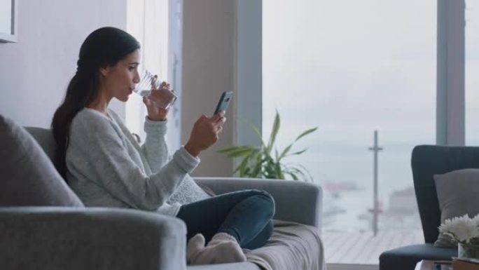 美丽的年轻女子使用智能手机在家沙发上放松浏览在线观看娱乐饮用水享受舒适的生活方式