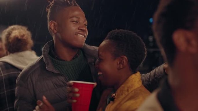 快乐的非洲裔美国夫妇亲吻在雨中跳舞，享受夜晚热闹的屋顶派对庆祝活动