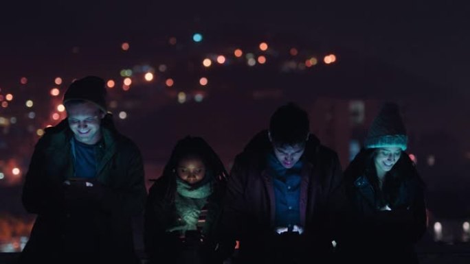 一群朋友晚上在屋顶上放松，使用智能手机技术浏览社交媒体在线聊天，享受周末聚会