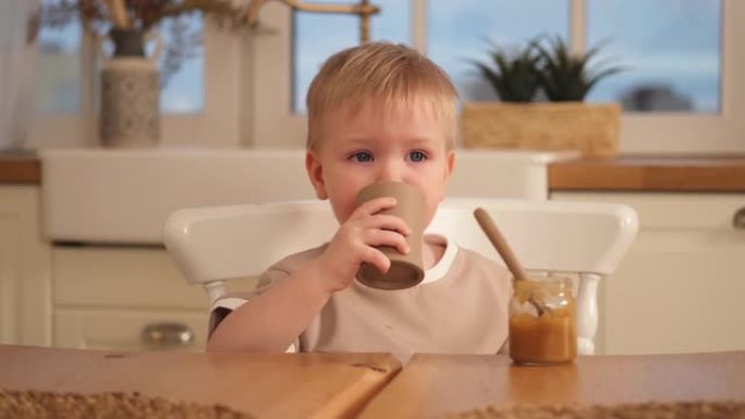 家庭幸福。男婴在厨房里喂自己。小男孩喝水，吃健康的食物。孩子拿着杯子自学吃饭。自食。