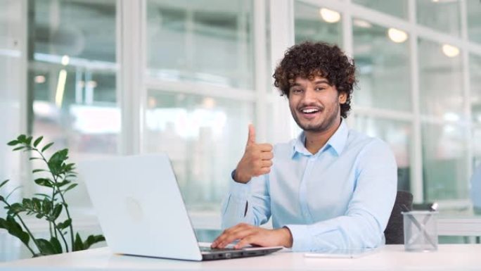 年轻的微笑工人坐在现代办公室的办公桌前，在笔记本电脑上工作时竖起大拇指。成功的正面男人在衬衫上打手势