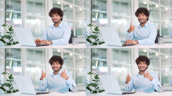年轻的微笑工人坐在现代办公室的办公桌前，在笔记本电脑上工作时竖起大拇指。成功的正面男人在衬衫上打手势