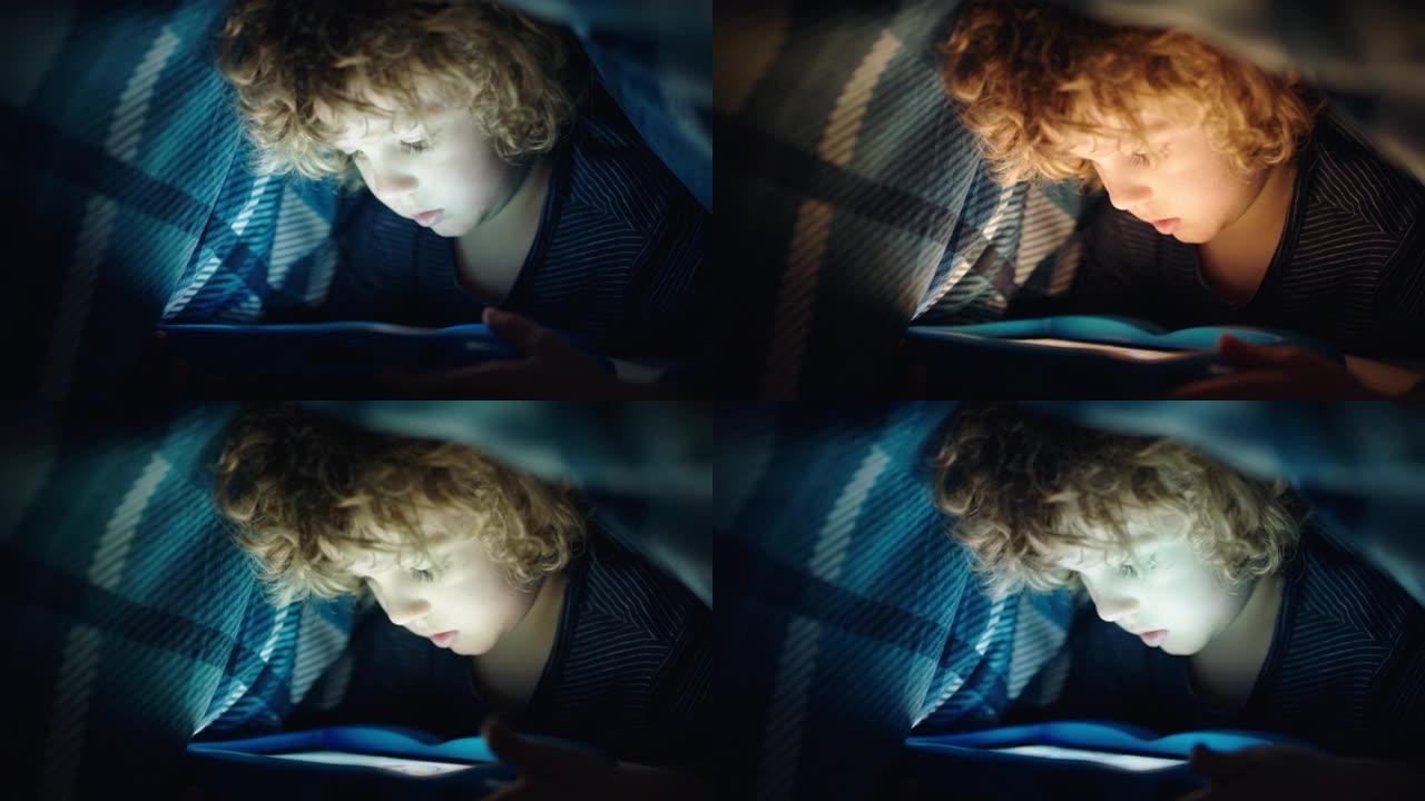 快乐的小男孩在毯子下使用数字平板电脑享受触摸屏技术的学习，在睡觉时玩游戏玩得开心