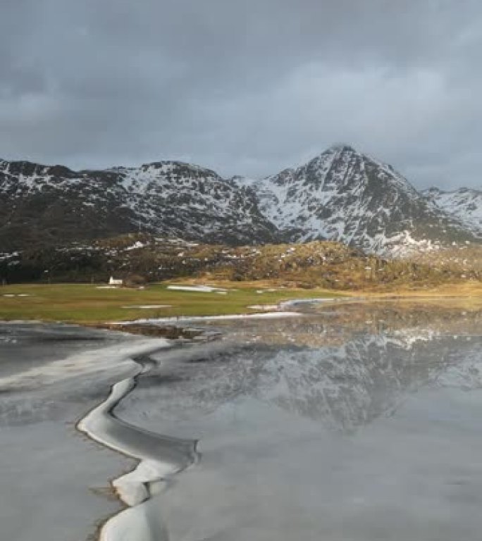 在寒冷的晴天，天空和云密布，美丽的白色冬季仙境全景，雪山山顶映衬在水晶般清澈的冰湖中。挪威峡湾山。
