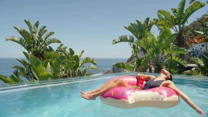 美丽的女人在粉红色的游泳管上放松漂浮在游泳池里喝果汁晒日光浴温暖阳光明媚的日子在豪华酒店度假村享受暑