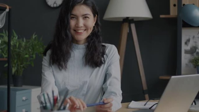 迷人的亚洲女性企业主微笑着坐在办公室的办公桌前的慢动作肖像