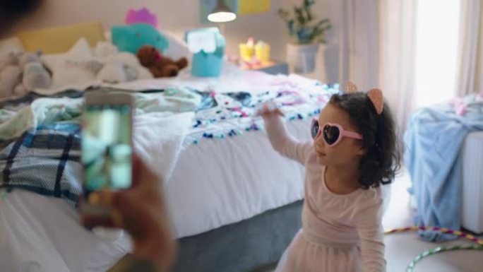 快乐的小女孩在卧室里玩装扮跳舞，母亲用智能手机在社交媒体上分享照片