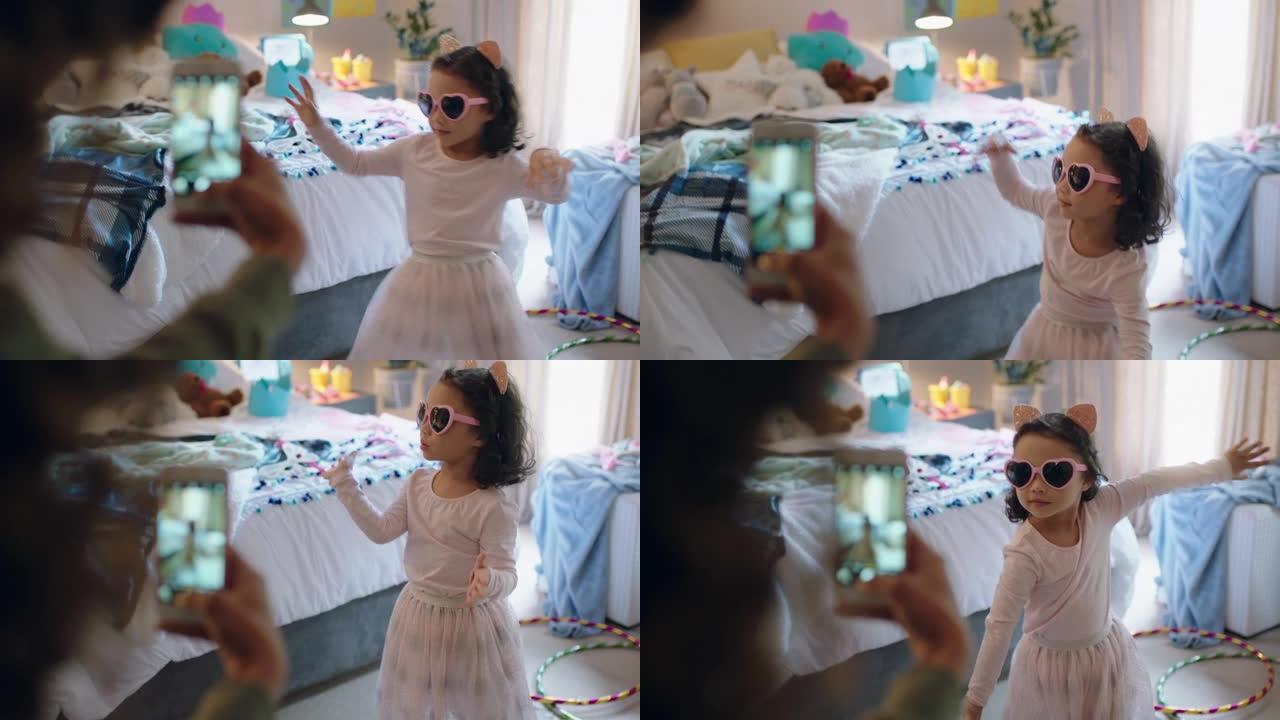 快乐的小女孩在卧室里玩装扮跳舞，母亲用智能手机在社交媒体上分享照片