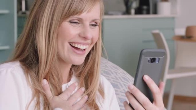 美丽的金发女人使用智能手机进行视频聊天，微笑着看起来很兴奋，享受着在家里用手机与朋友聊天的好消息