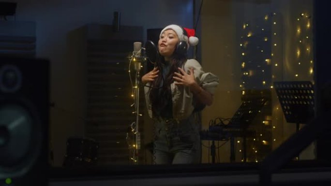 一位专业的亚洲女歌手在录音室为她的圣诞节专辑录制歌曲的肖像。美丽的年轻艺术家，戴着圣诞老人的帽子，喜