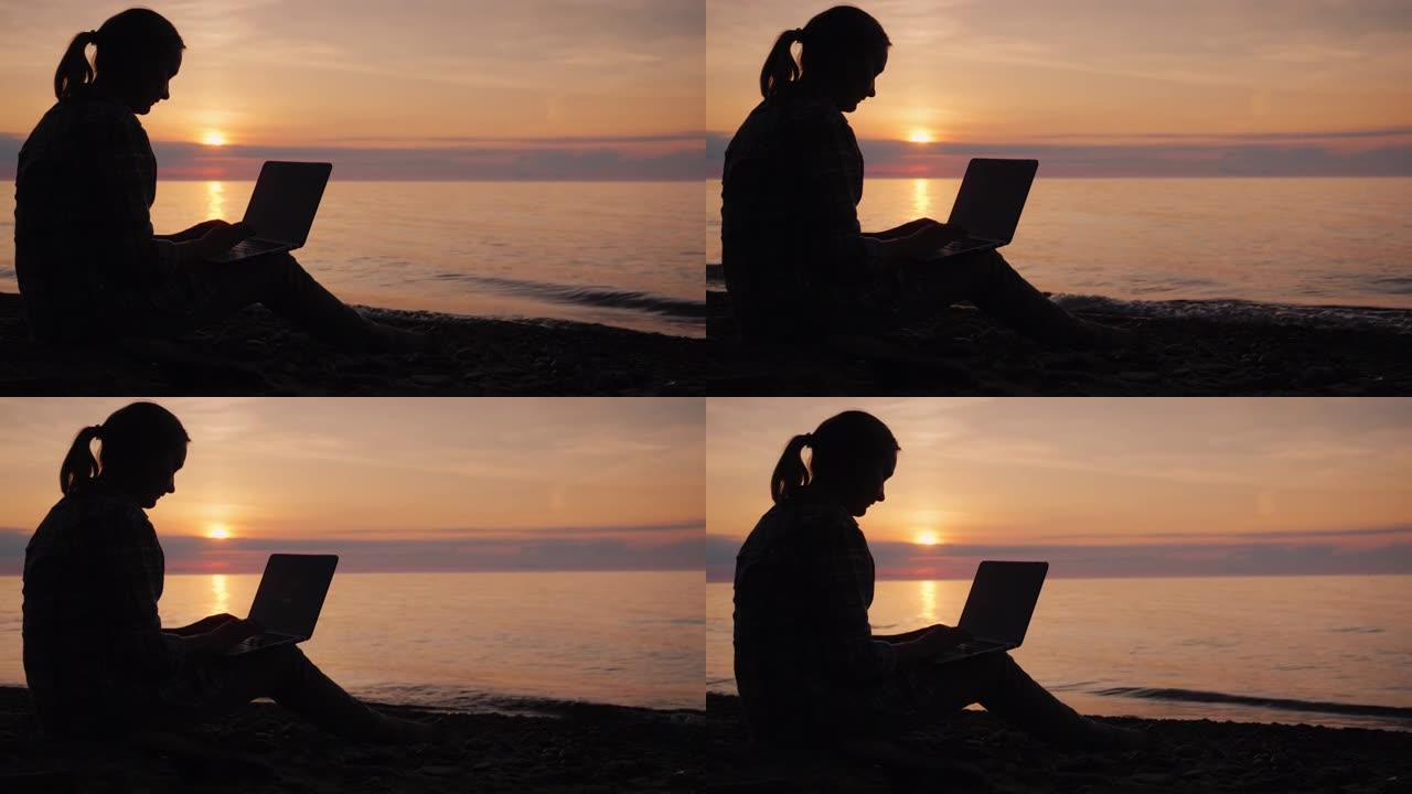 日落时在海边用笔记本电脑工作的女人的侧影