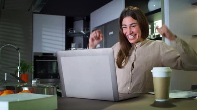 年轻成功的女商人在家厨房工作时在笔记本电脑上查看电子邮件的肖像。兴奋的女员工庆祝职业生涯中的成功交易