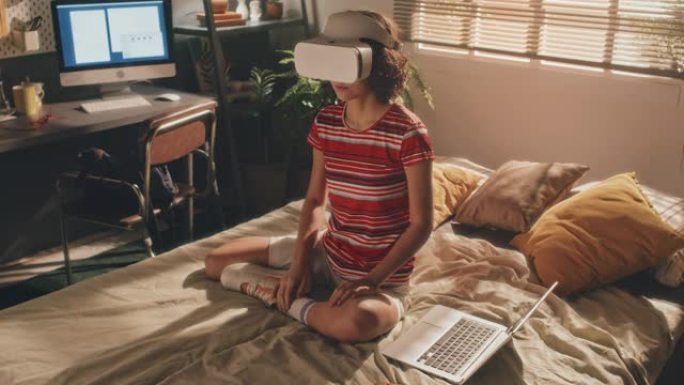戴着虚拟现实耳机的年轻卷发女孩坐在阳光充足的卧室的床上