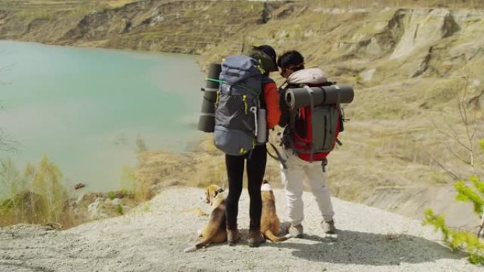 两名带着背包的游客带着两只狗在湖岸看相机照片