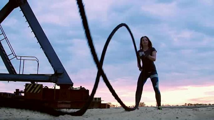 年轻，运动，女人，在厚厚的运动绳索的帮助下进行力量练习。黎明时分，在沙质采石场的海滩上，在阳光下，靠