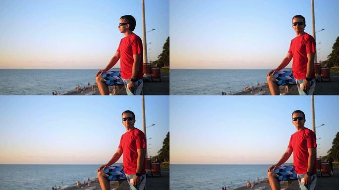 日落时，戴着墨镜的年轻英俊男子在海边放松。他欣赏日落和水。转过头看着相机后
