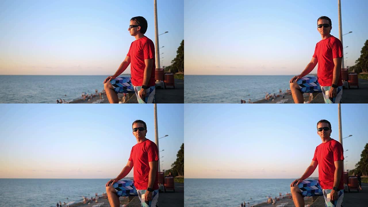 日落时，戴着墨镜的年轻英俊男子在海边放松。他欣赏日落和水。转过头看着相机后