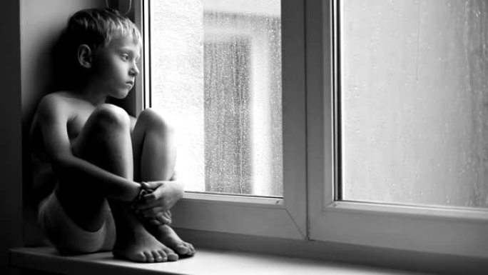 一个悲伤的男孩在倾盆大雨时坐在窗台上，无法离开公寓的黑白镜头。