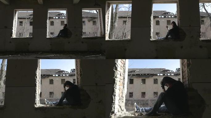 蒙面沮丧的年轻人坐在一栋废弃建筑的窗框上