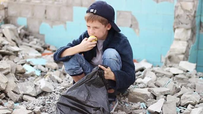 无家可归的孤儿男孩在房子的废墟中寻找包装中的食物并吃苹果