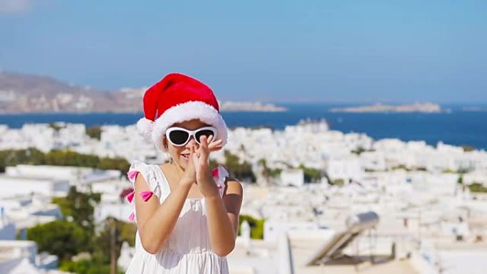 米科诺斯户外背景的红色圣诞老人帽子里有趣的小女孩。希腊圣诞节假期，孩子在典型的希腊传统村庄的街道上，