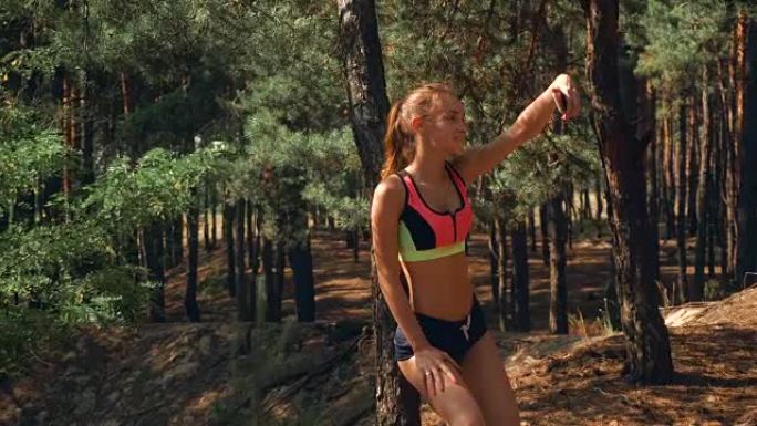 腹部有弹性的运动女孩站在树林里，看着你的手机