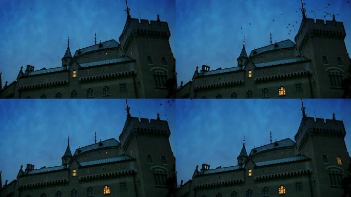 中世纪城堡场景: 随着黄昏的降临，灯光逐渐出现在窗户中。一群鸟从屋顶上方经过。