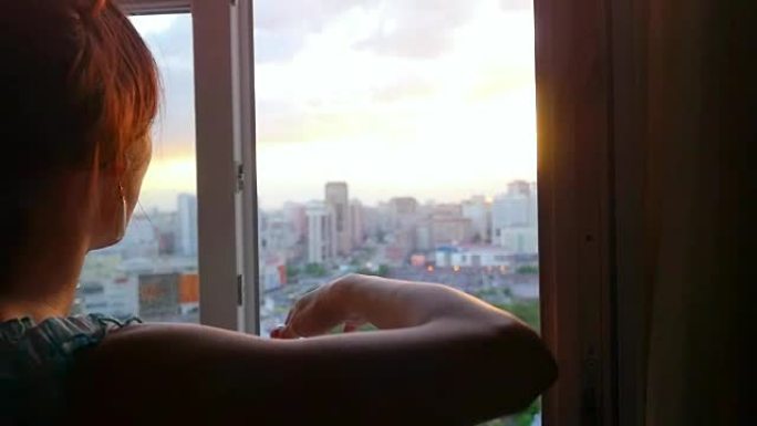 年轻女子打开窗帘，在美丽的日落时分，从俯视到城市从窗户到城市